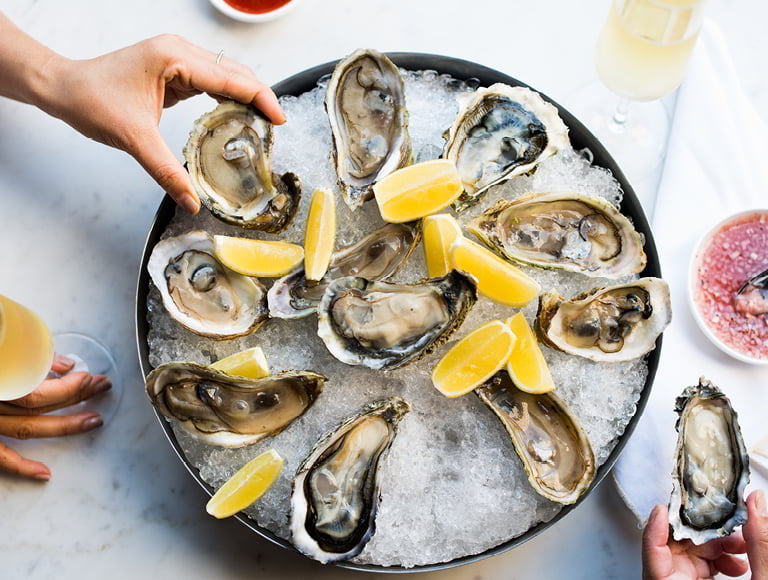Hands lifting an oyster from a tray of iced oysters arranged with lemon wedges beside glasses of white wine and a bowl of mignonette.