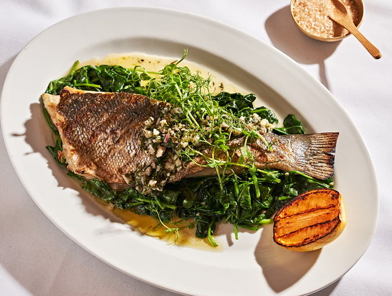 Whole roasted fish topped with chopped herbs and onions served over sautéed greens with a charred lemon half on a white plate.